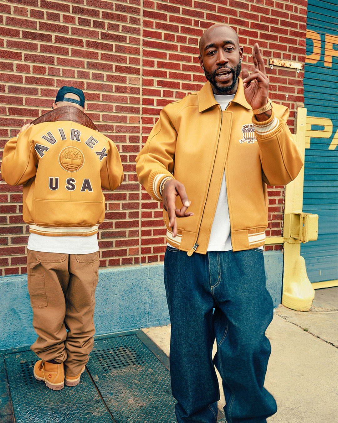 Two men wearing mustard yellow bomber jackets and baggy pants against a brick wall.