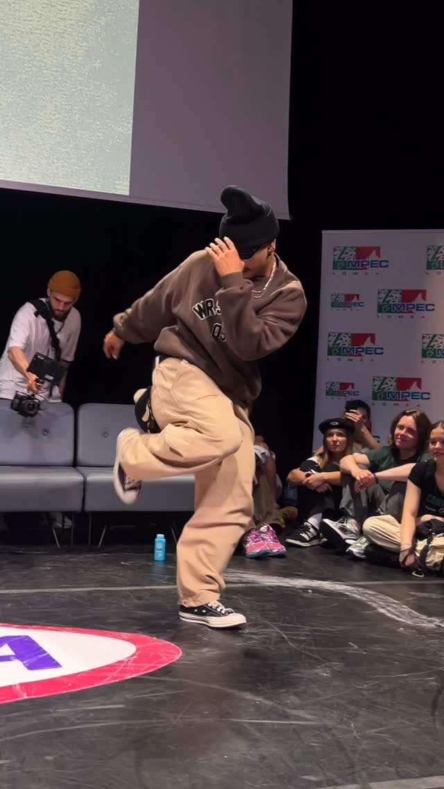 A dancer wearing baggy tan pants and a brown sweatshirt performs on a stage.