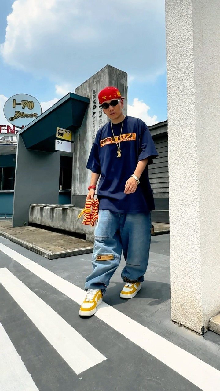 A young man wearing baggy jeans, a large graphic t-shirt, and a red cap posing on a crosswalk.