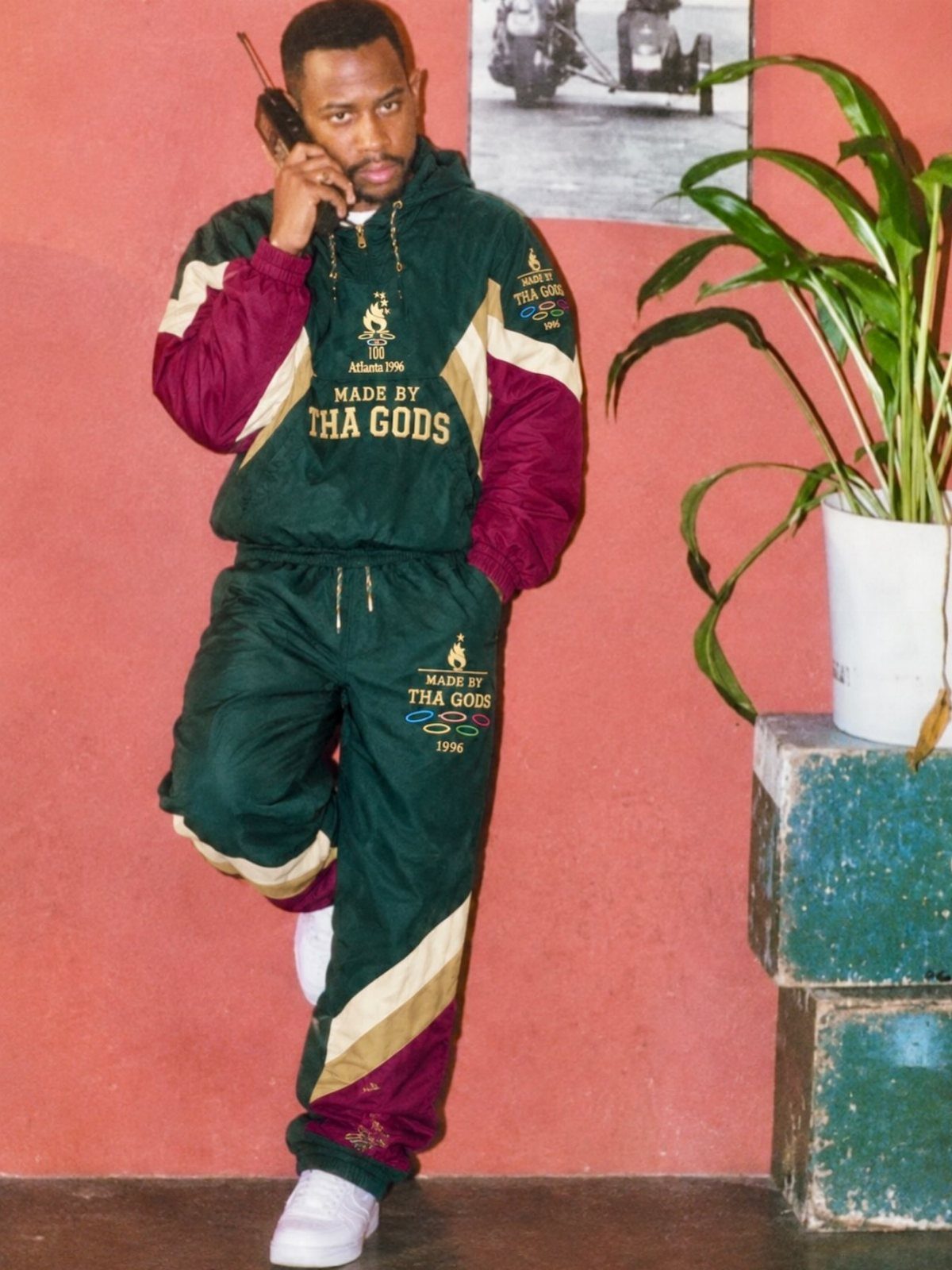 Man in a green and burgundy tracksuit holding a walkie-talkie against a pink wall.