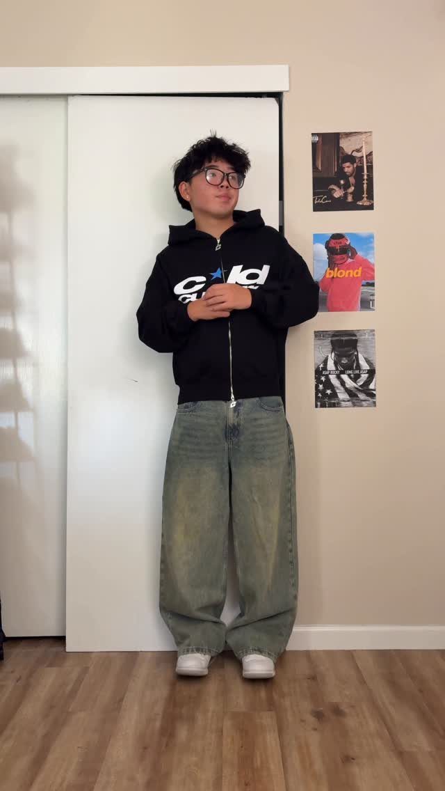 A young man posing in a black graphic hoodie and baggy denim jeans.