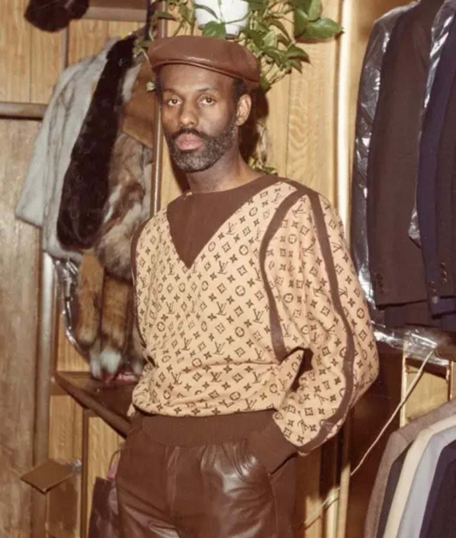 A man wearing a brown beret, patterned sweater, and leather pants standing in a clothing shop.