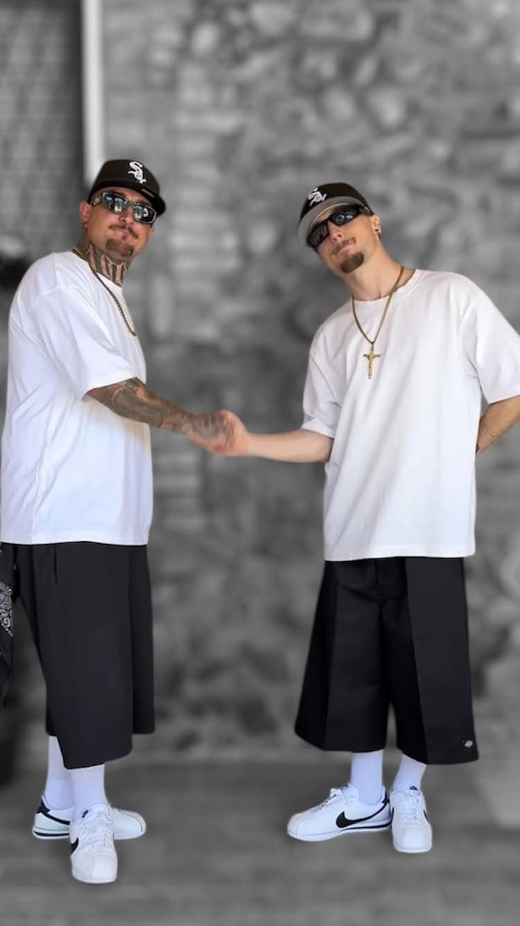 Two men in oversized white shirts, black shorts, and baseball caps posing together.