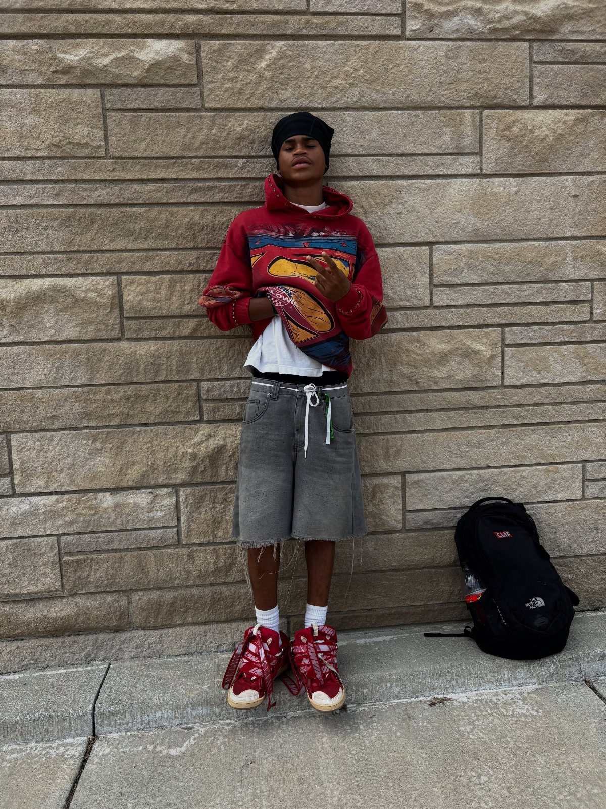 Person in streetwear standing against a stone wall wearing a red hoodie and grey shorts.