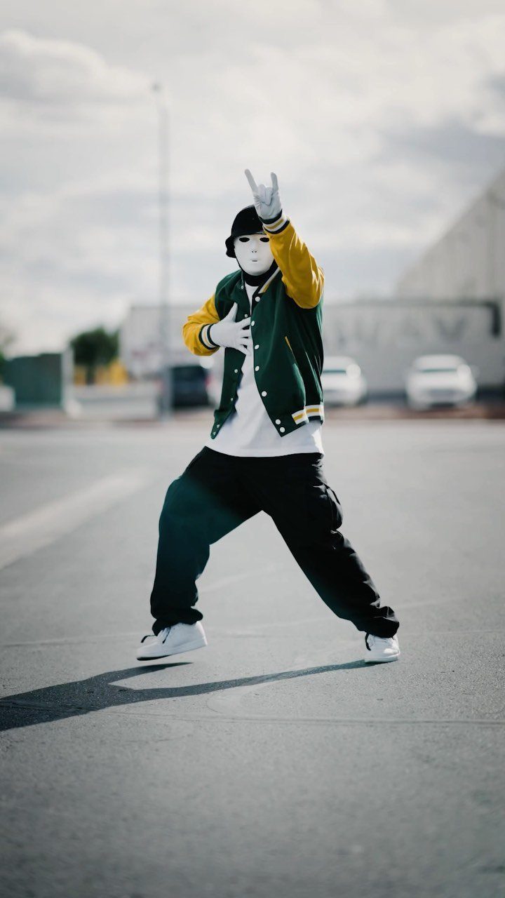 A person in hip hop attire including a varsity jacket and mask posing on a street.