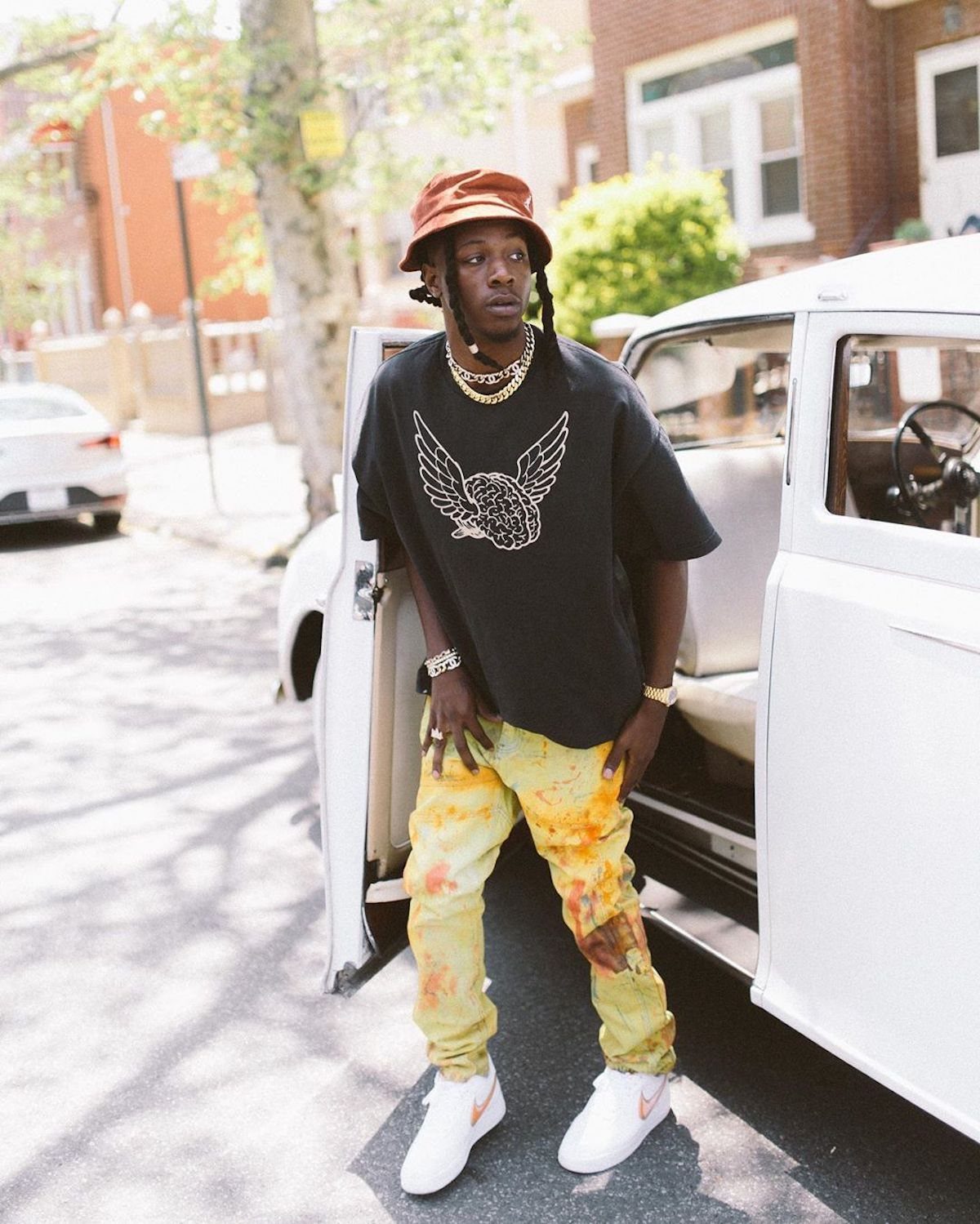 A man in an orange bucket hat and black graphic t-shirt posing by a white car.