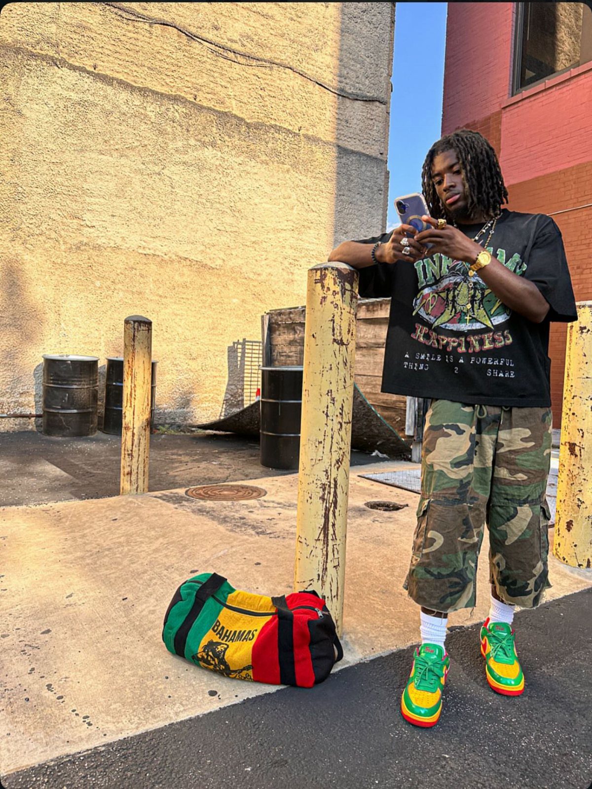 A man in a graphic tee, camo shorts, and bright sneakers poses in an urban alleyway.