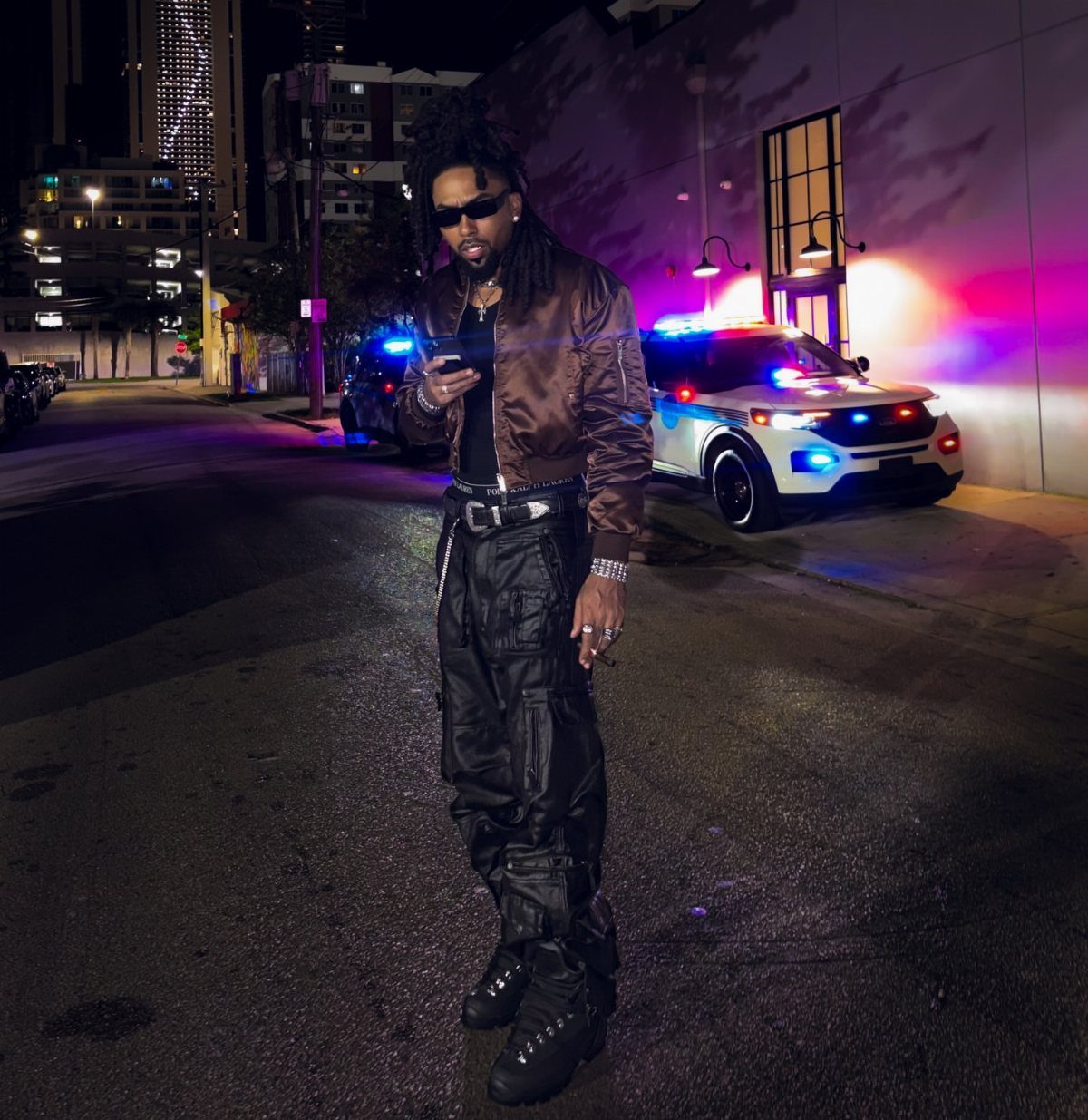 Man in a brown bomber jacket and black cargo pants standing on a city street at night.