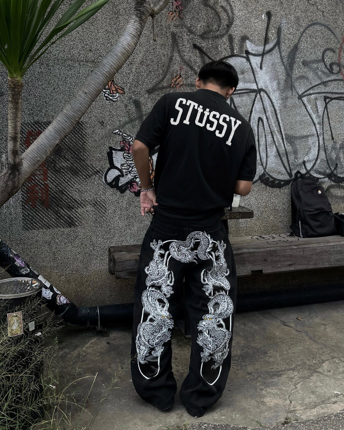 A person wearing a black Stussy shirt and dragon embroidered baggy pants standing by a graffiti wall.
