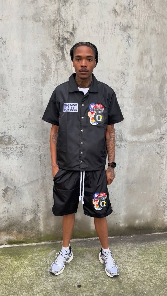 Man in a black graphic short-sleeved shirt and matching shorts standing against a concrete wall.