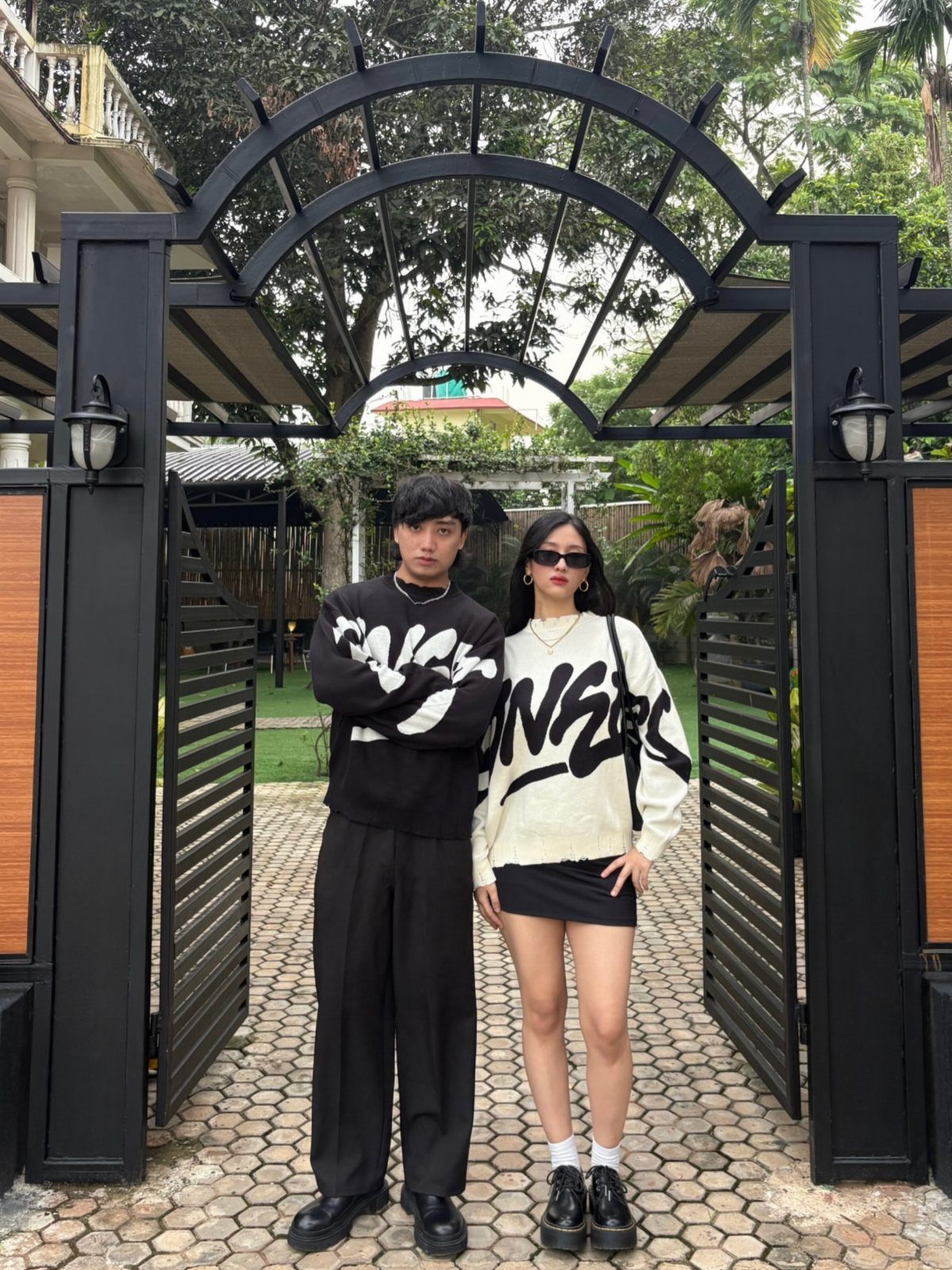 A young man and woman posing in stylish streetwear outfits in front of a black gate.