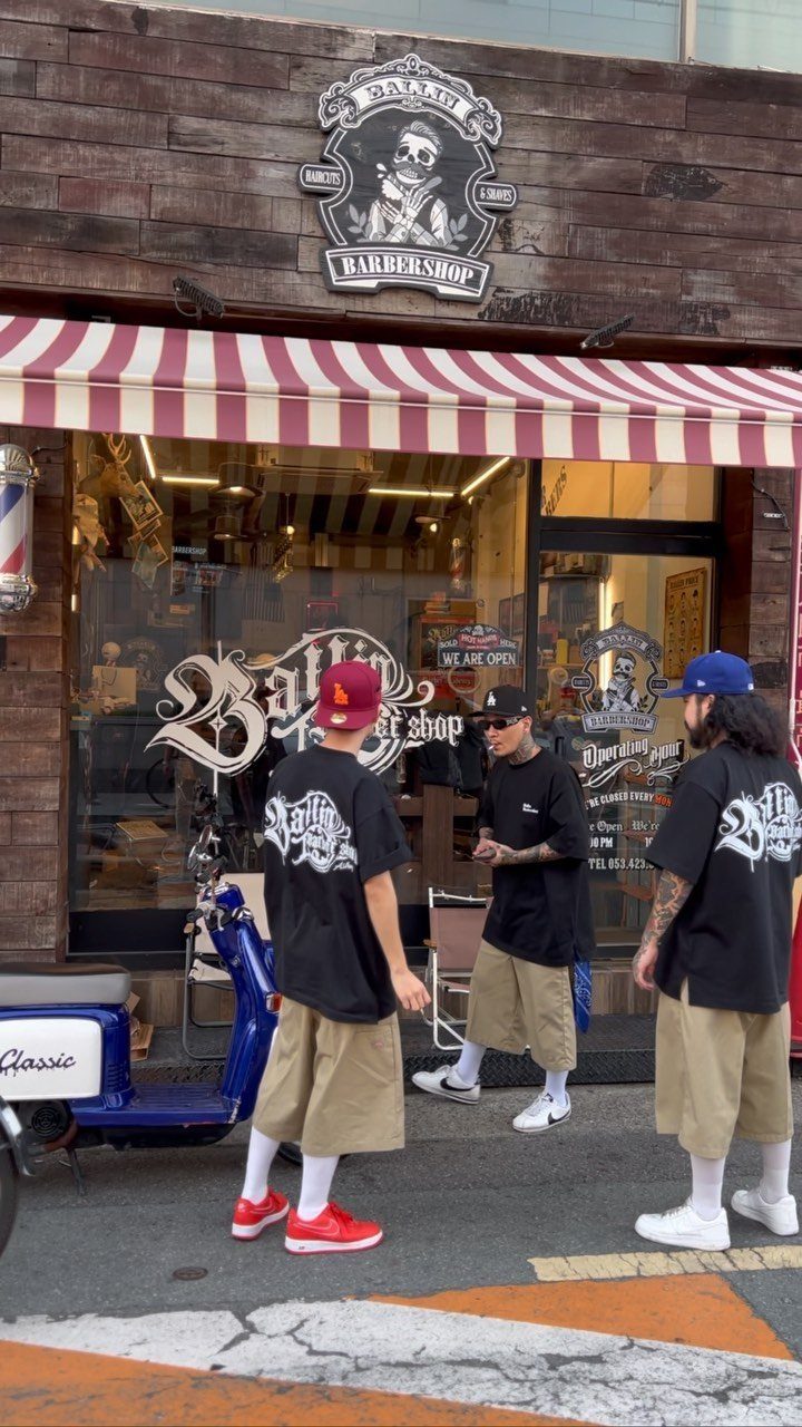 Three men in oversized graphic t-shirts and baggy shorts standing outside a barbershop.