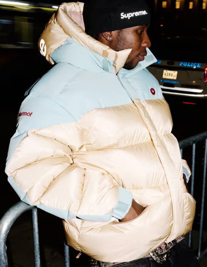 A man wearing a light blue and beige puffer jacket and a black Supreme beanie.