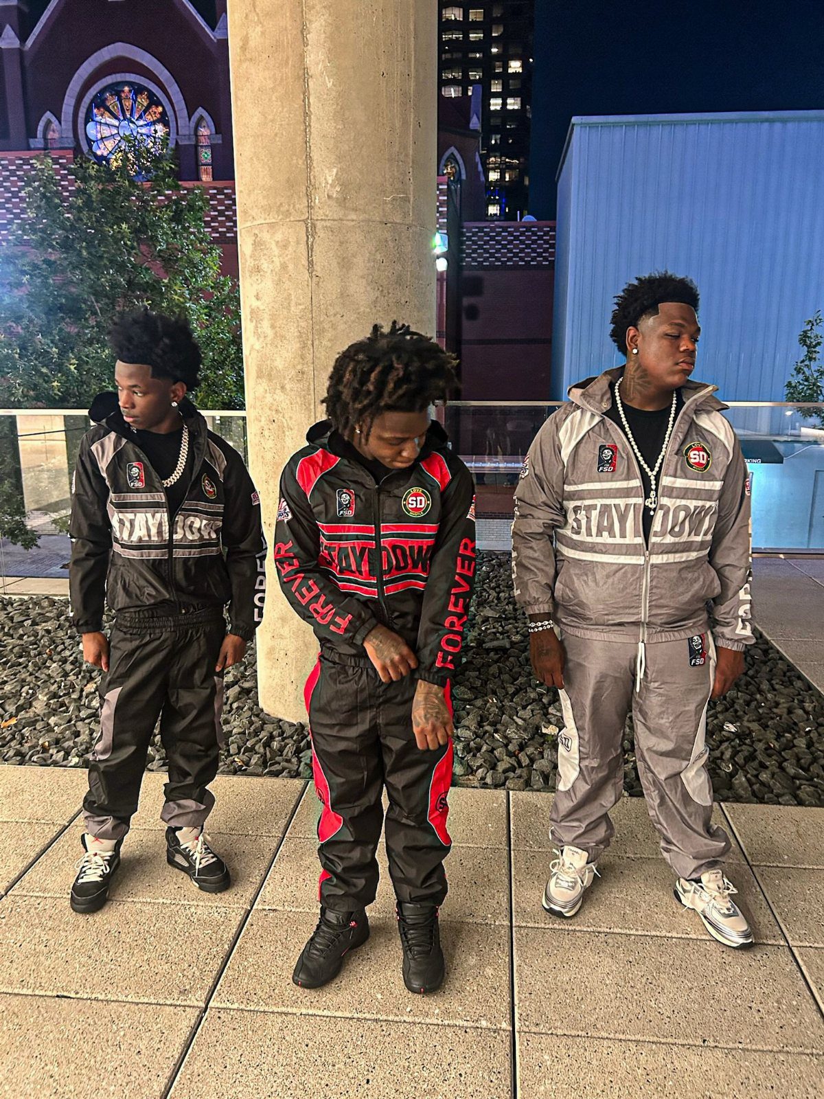 Three men standing outdoors at night wearing coordinated urban streetwear tracksuits with bold lettering.