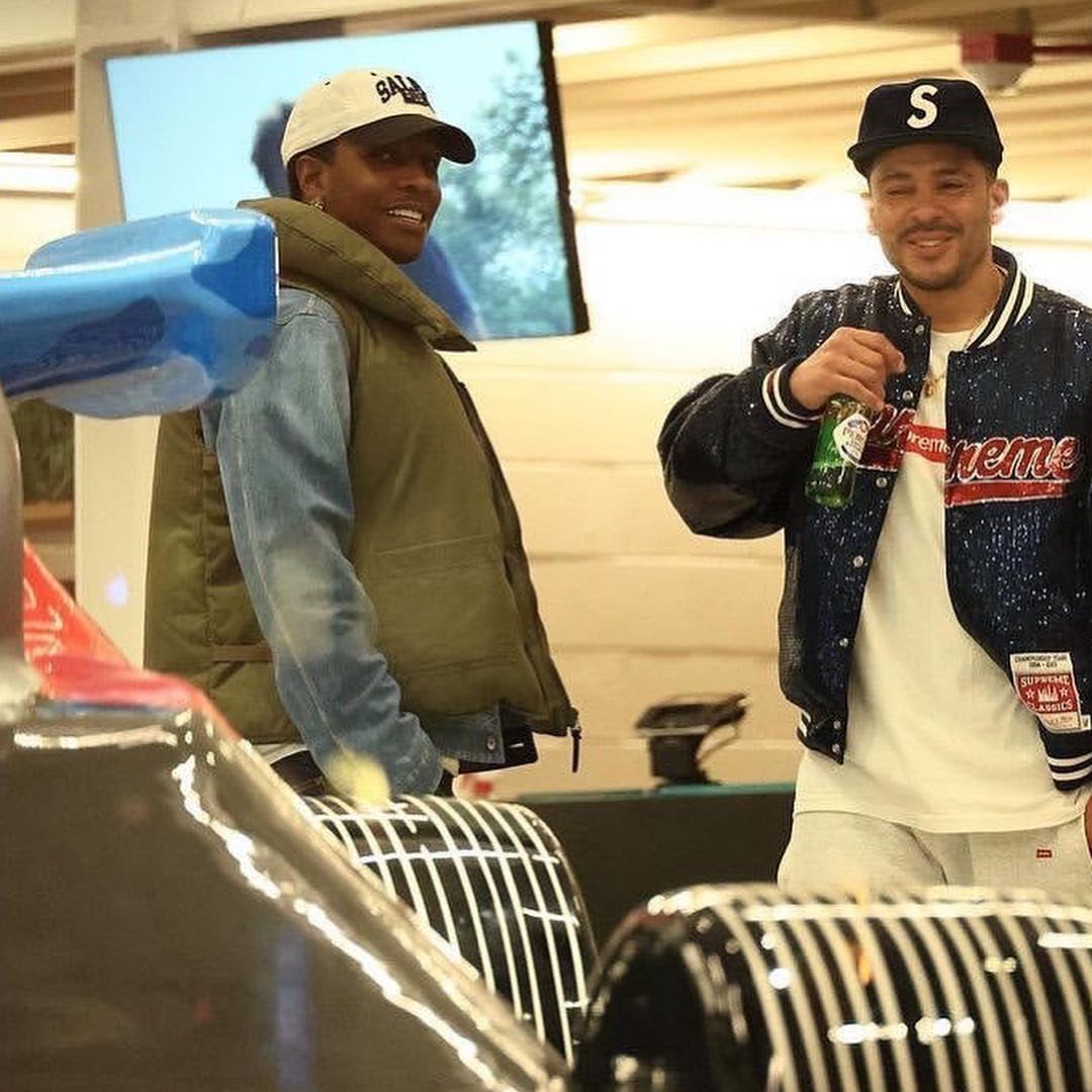 Two men posing in stylish streetwear near a car with a screen in the background.