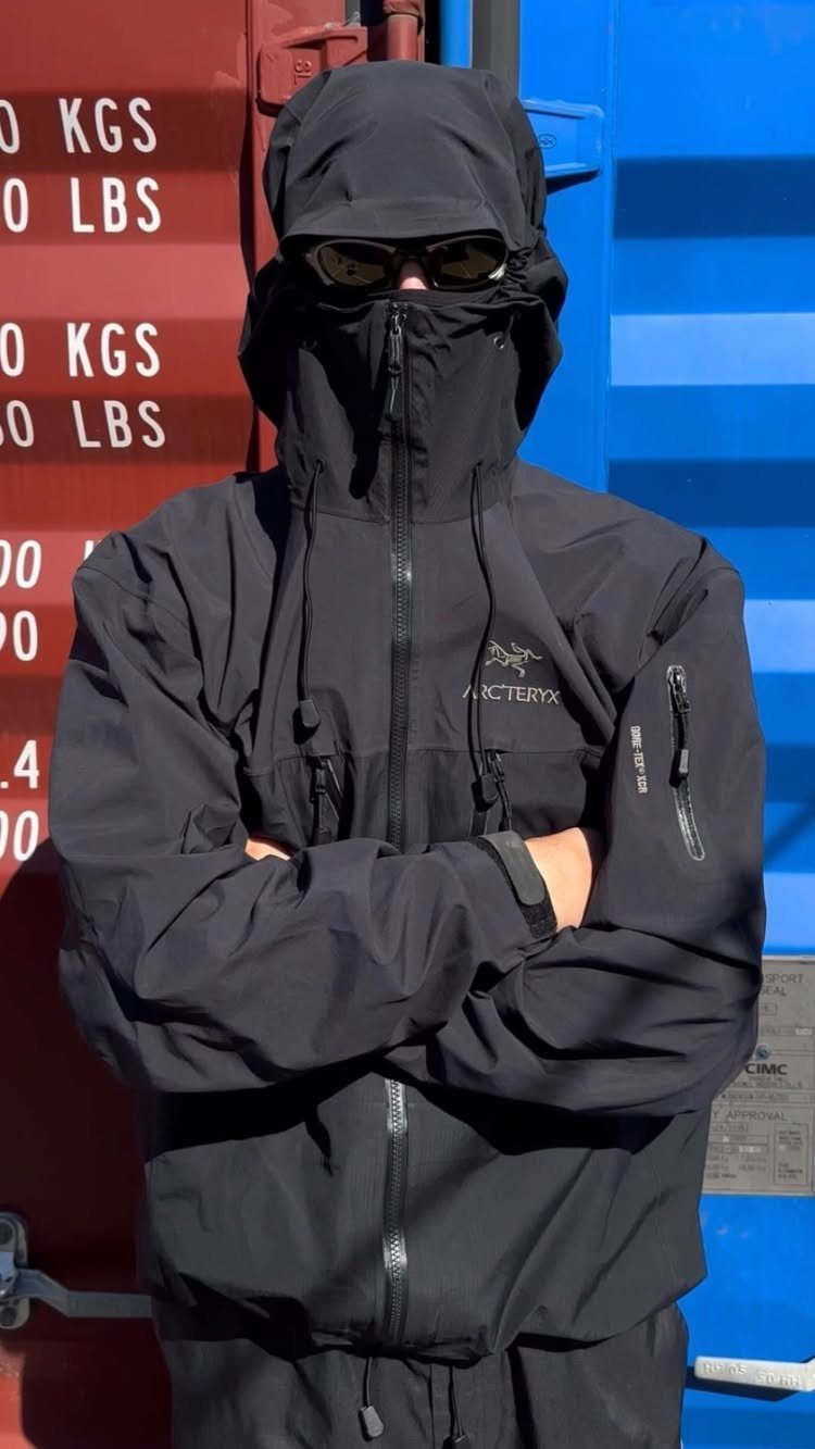 Man in black technical hooded jacket and sunglasses standing with arms crossed against a colorful background.
