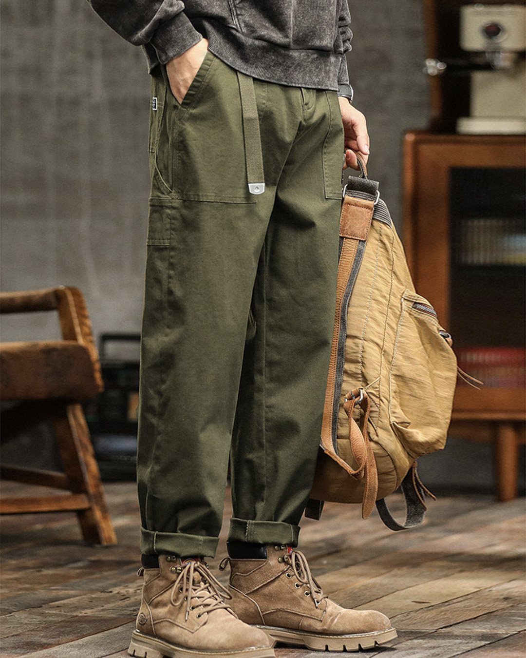 A person wearing olive green cargo pants, a grey sweatshirt, and tan boots while holding a backpack.