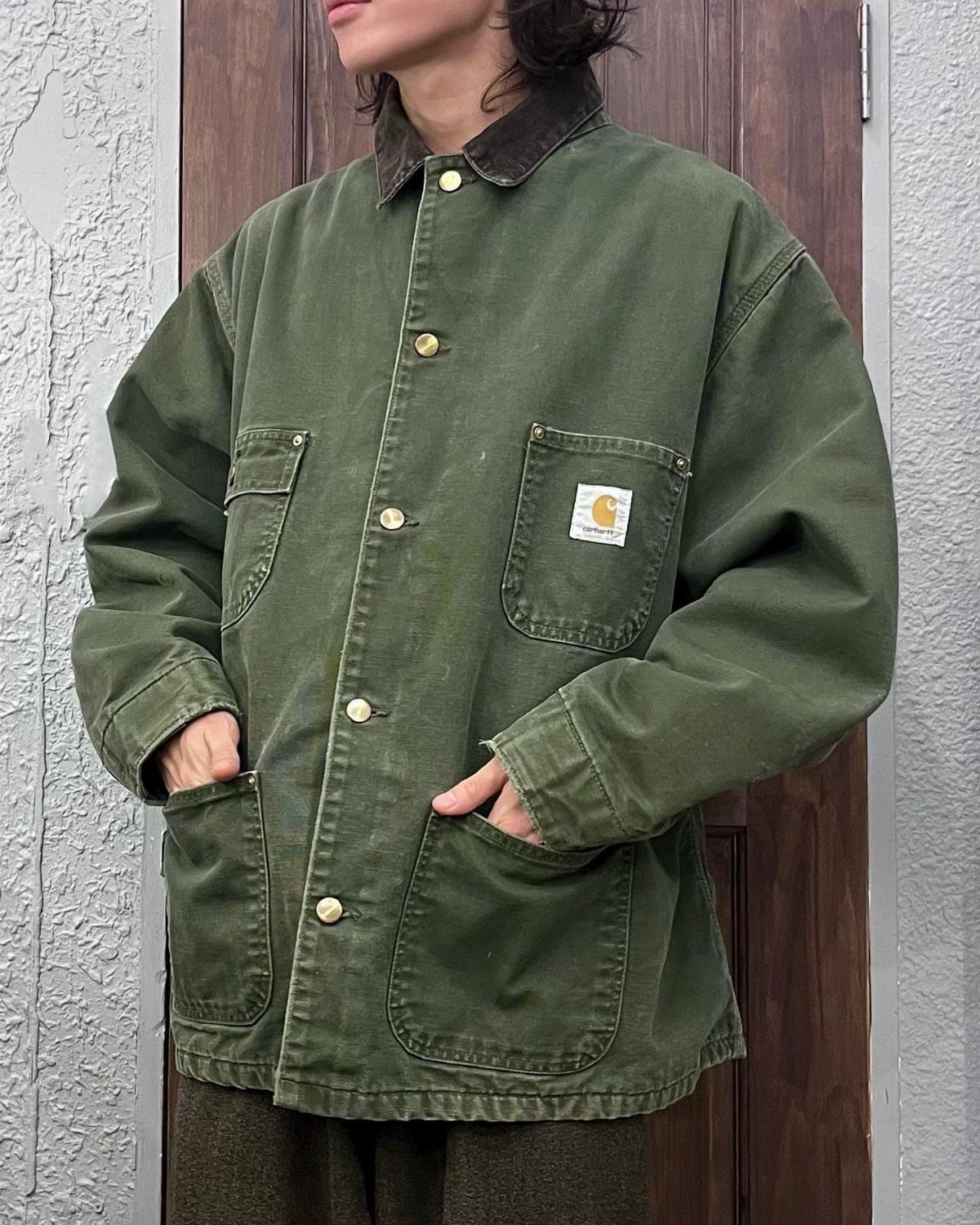 A person wearing an oversized olive green canvas chore coat with large patch pockets and brass buttons.