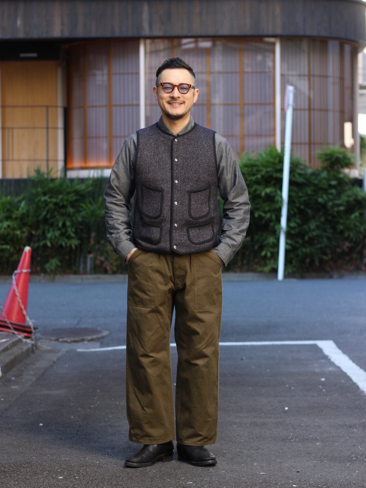 A man smiling while wearing a textured vest, grey shirt, and wide olive trousers on a city street.