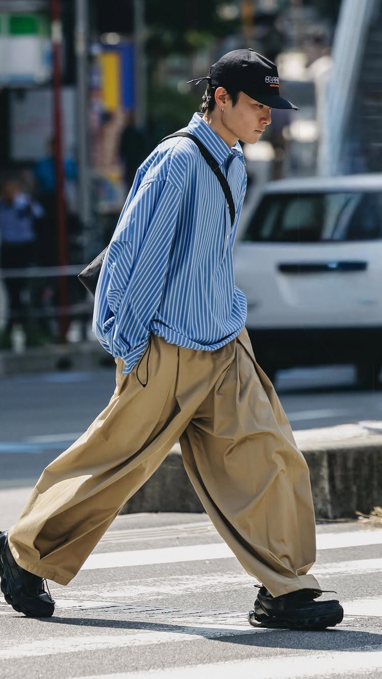Man walking in oversized blue striped shirt and wide tan trousers on a city street.