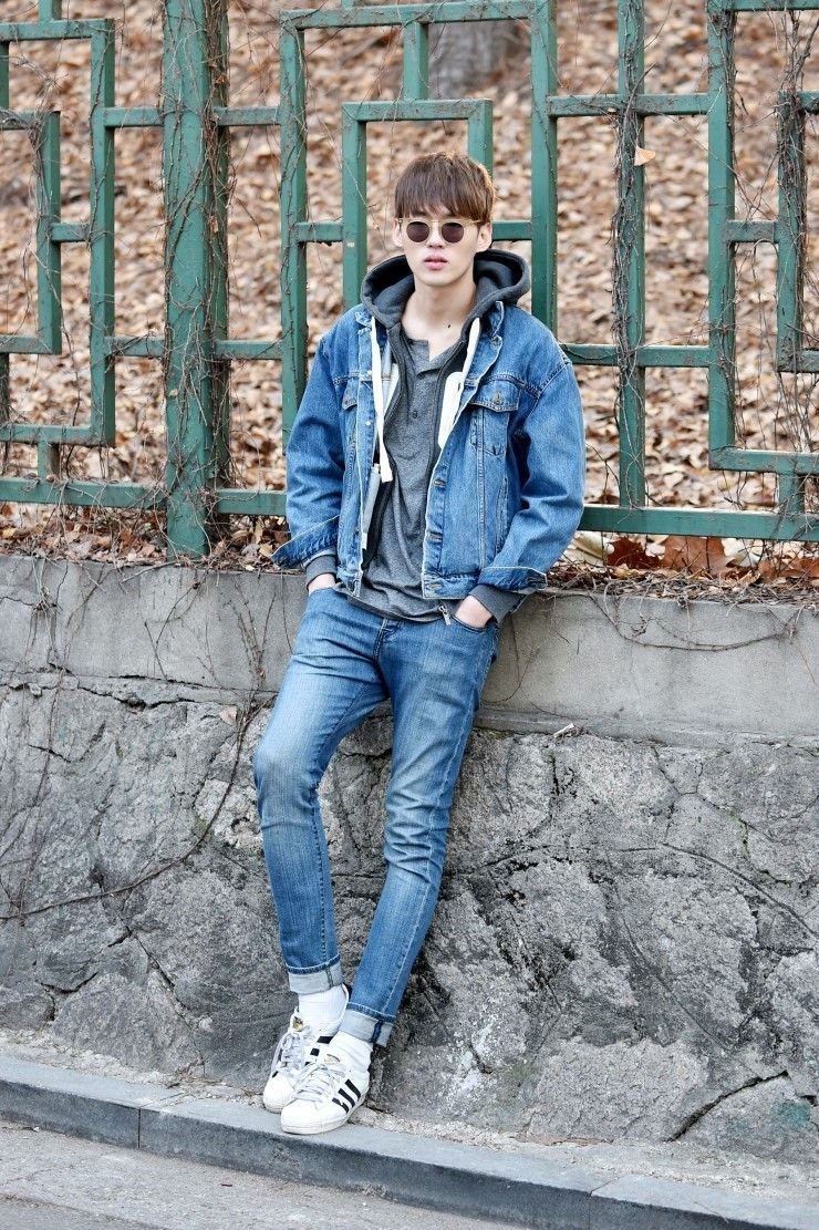 A young man in a denim jacket and hoodie leaning against a stone wall outdoors.