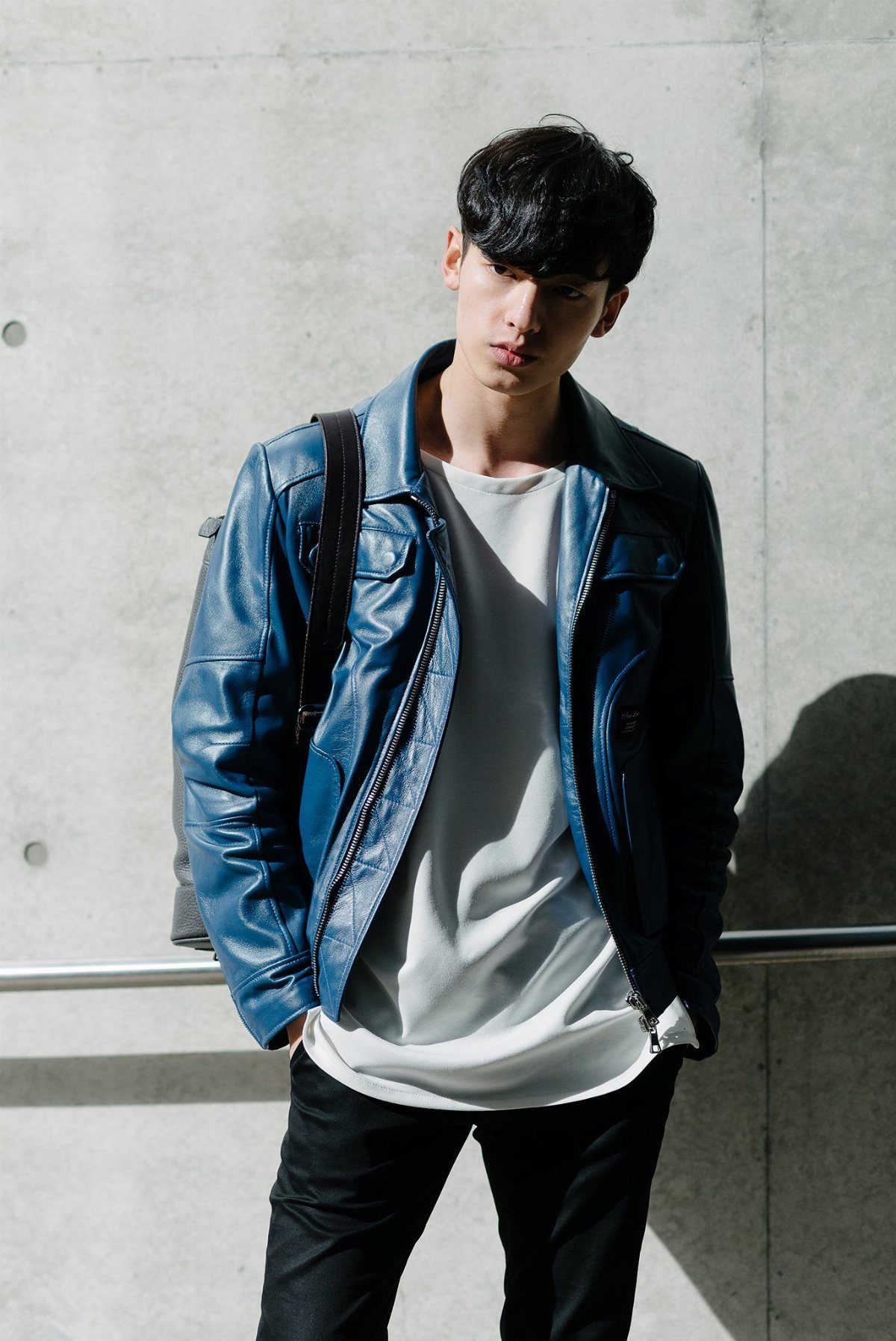 A young man posing in a blue leather jacket and white t-shirt against concrete.