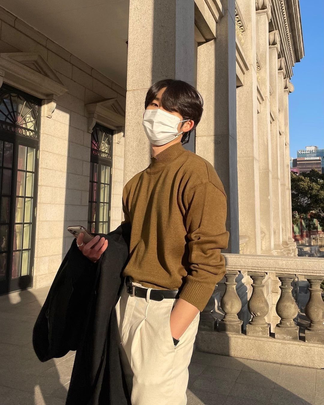 Man in a brown mock neck sweater and cream pleated trousers standing by stone columns.