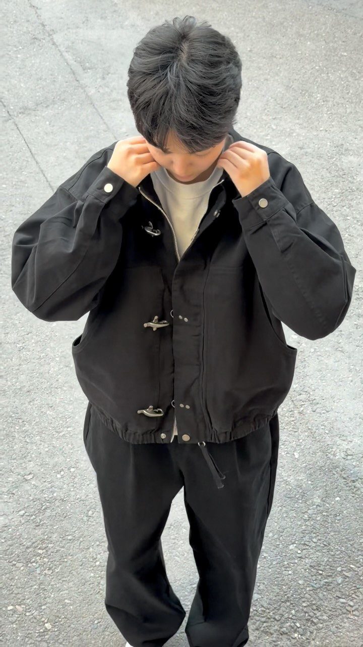 A man wearing a black utility jacket with toggle fasteners and black trousers on asphalt.