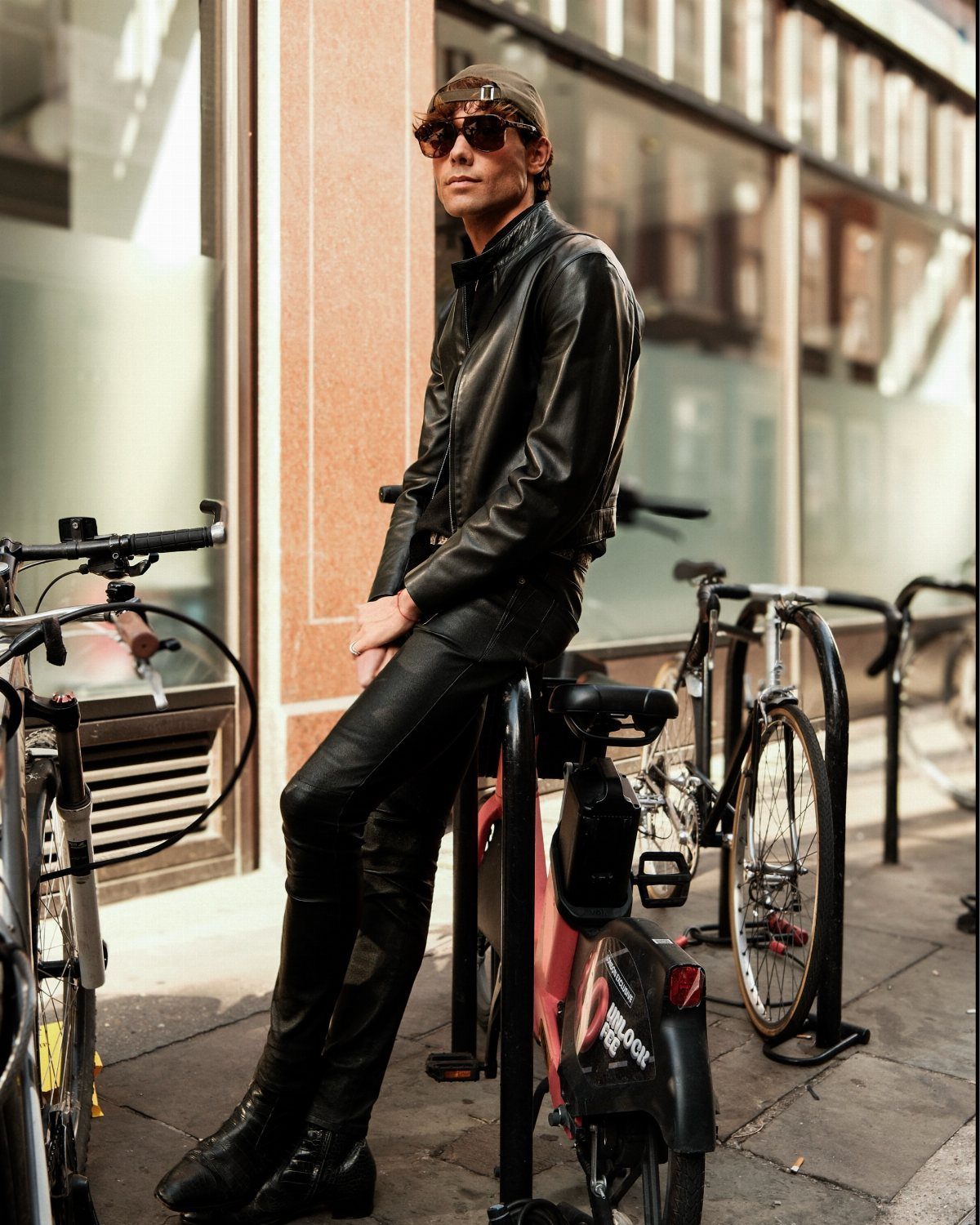 A man in a black leather outfit and sunglasses sits on a bike rack on a city street.