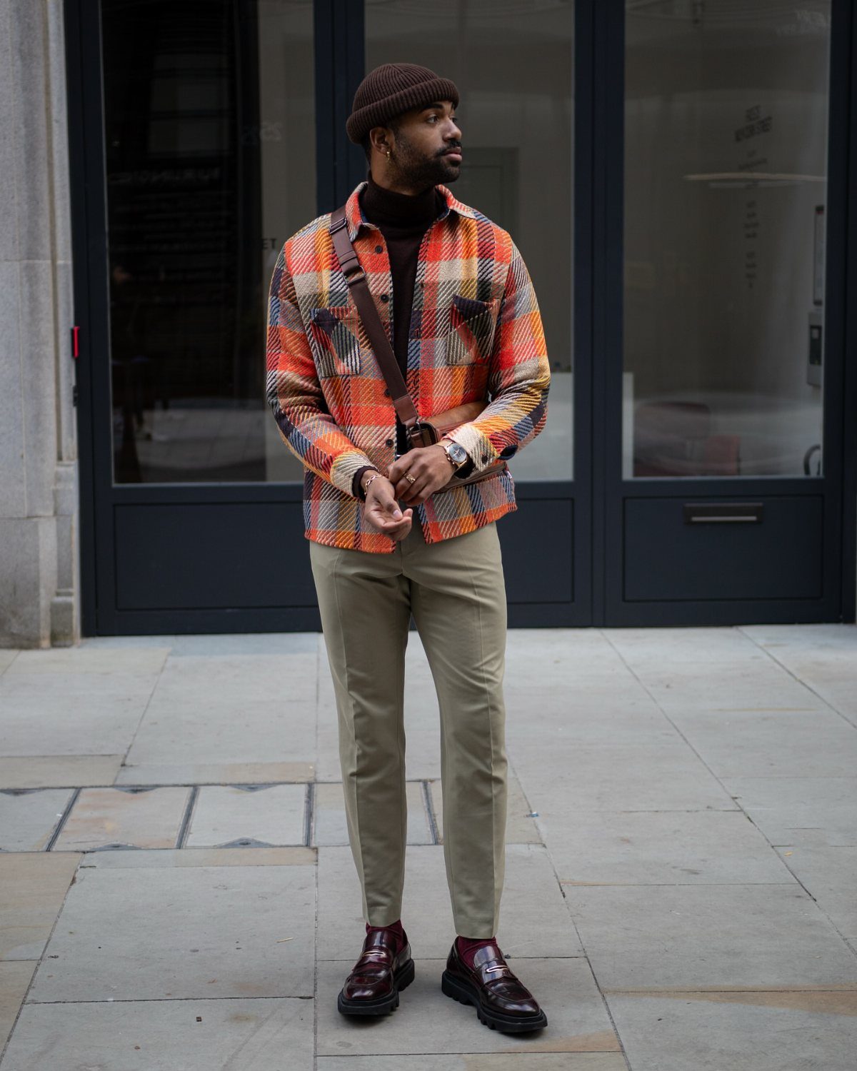Man in a colorful plaid shirt, black turtleneck, tan trousers, and burgundy loafers standing outdoors.