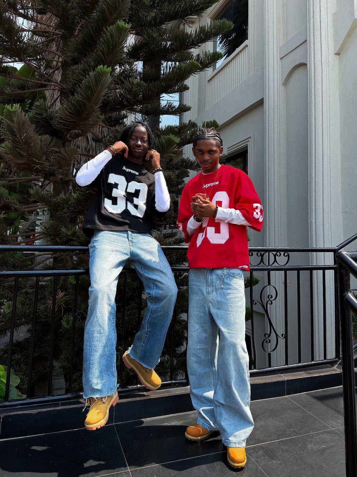 Two men posing in baggy denim jeans and oversized graphic long-sleeve shirts on a balcony.