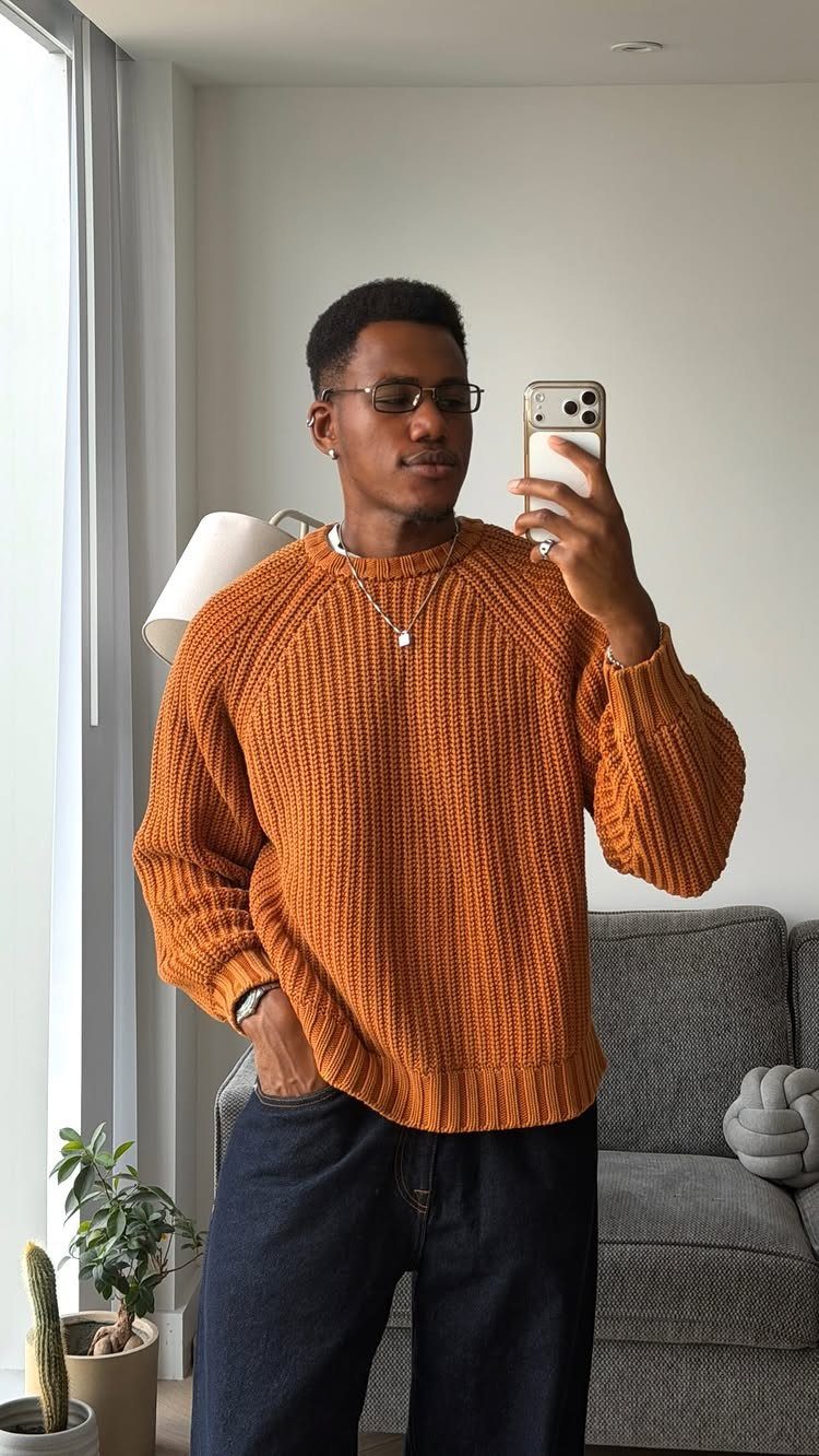Man in an orange textured knit sweater and dark jeans taking a mirror selfie.
