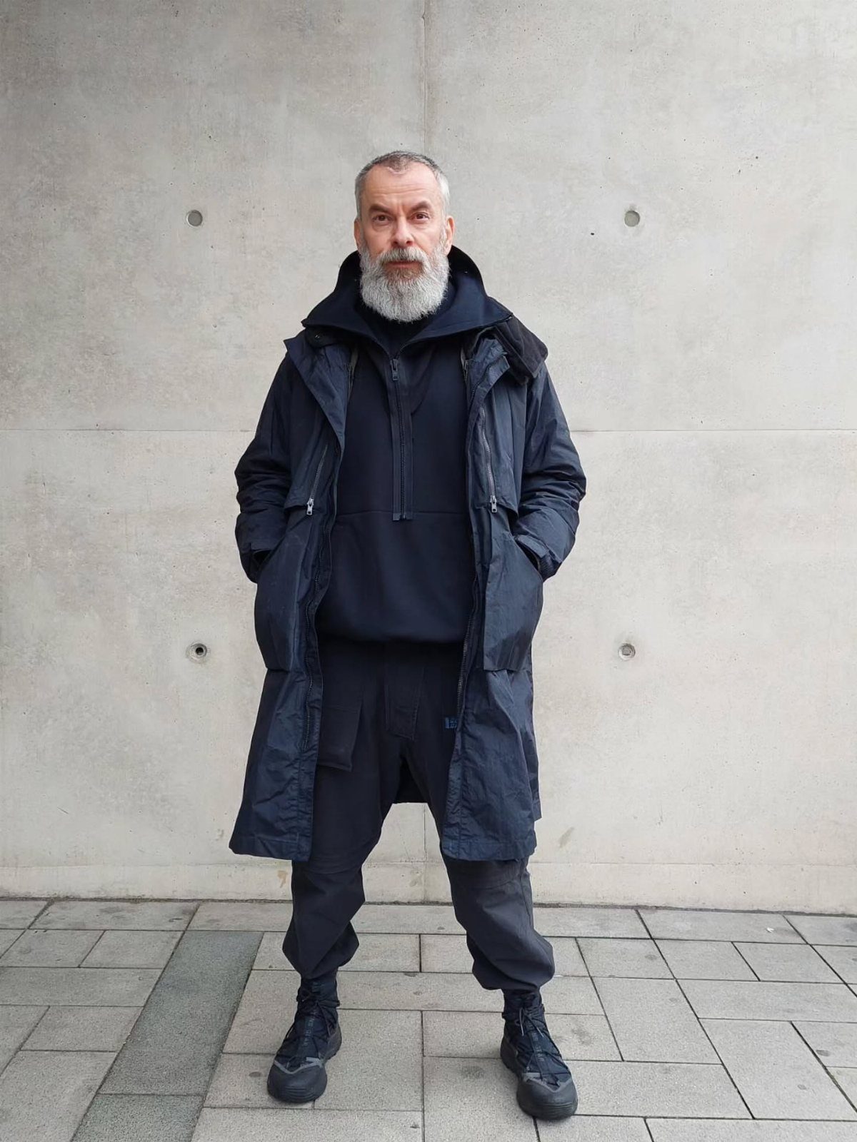 Man in black technical streetwear standing against a grey concrete wall.
