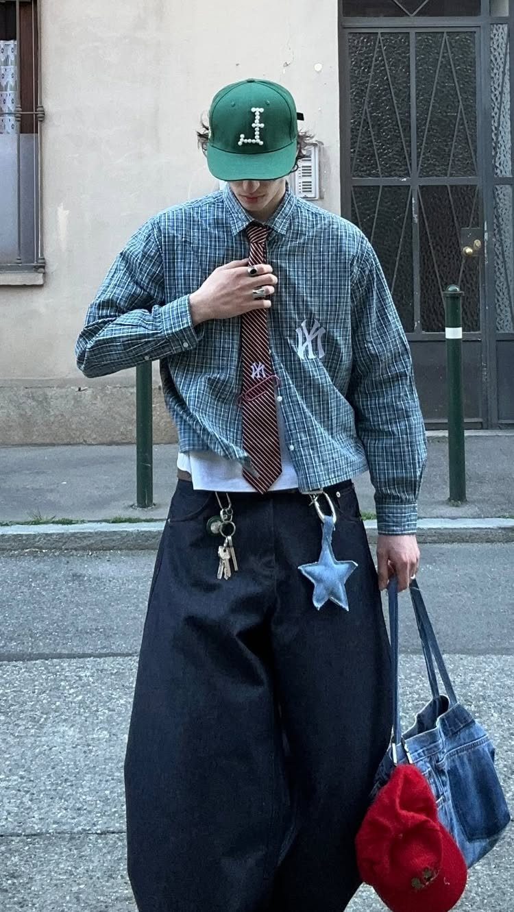 A person wearing a checkered shirt, brown tie, and wide black pants while holding a denim bag.