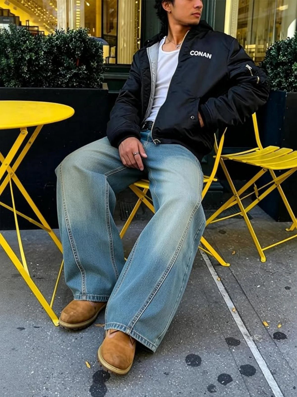 A man wearing a black bomber jacket, wide-leg blue jeans, and tan suede shoes sitting outdoors.