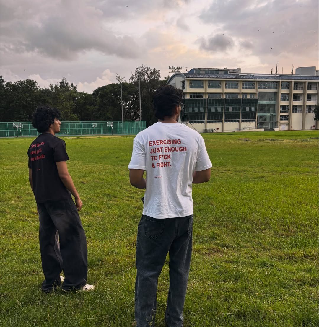 Two men standing in a field, one wearing a white graphic t-shirt with red text.