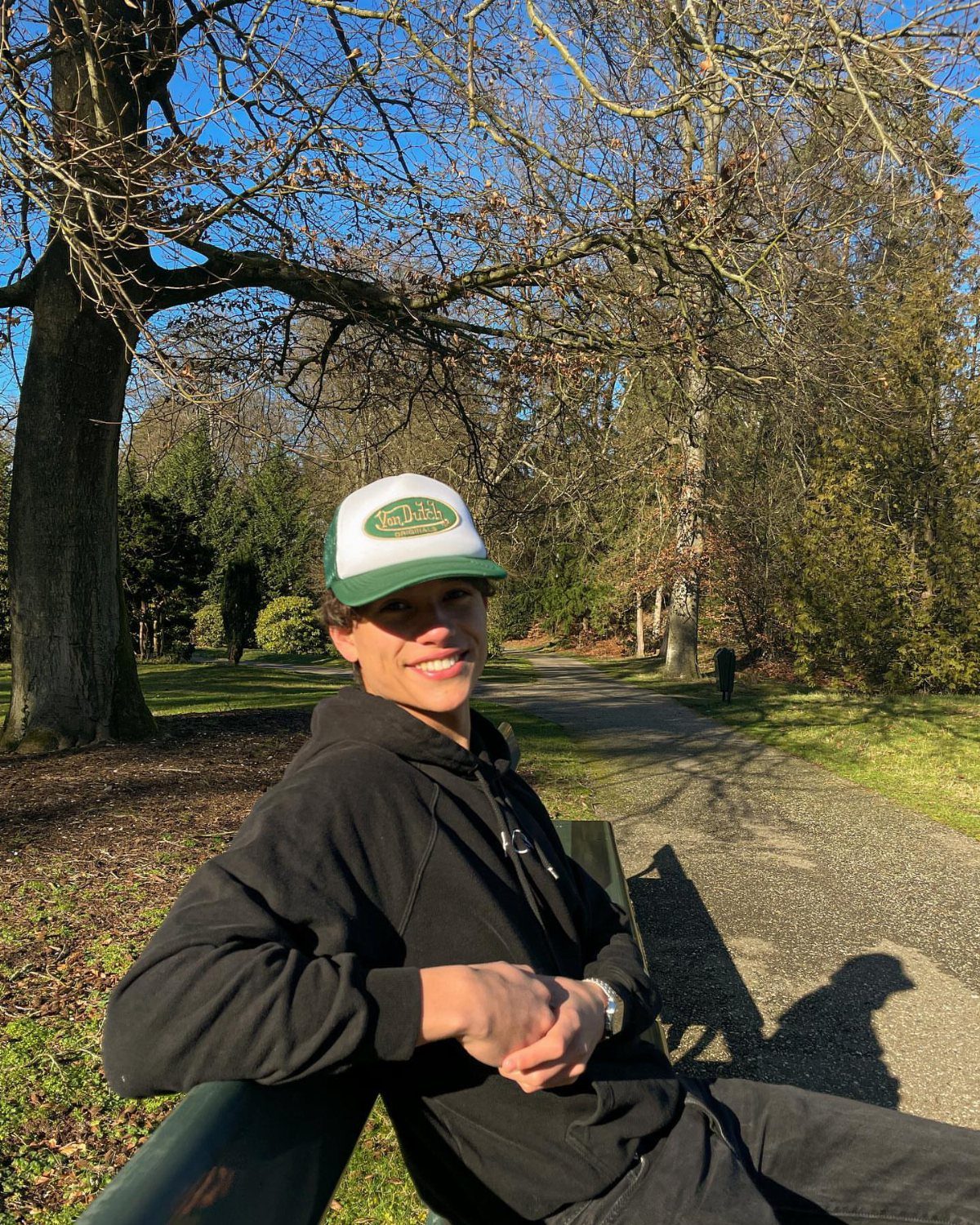A smiling man wearing a black hoodie and a green trucker hat sitting on a park bench.