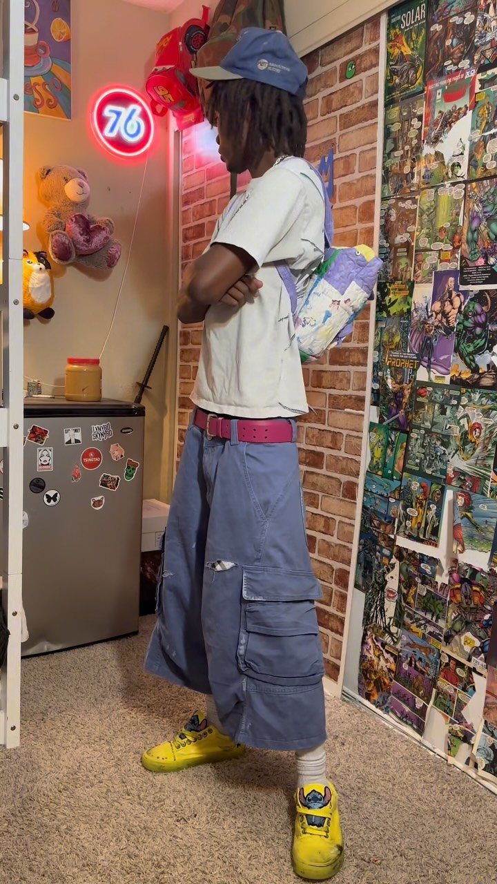 A young man posing in oversized blue cargo shorts, white t-shirt, and bright yellow sneakers.