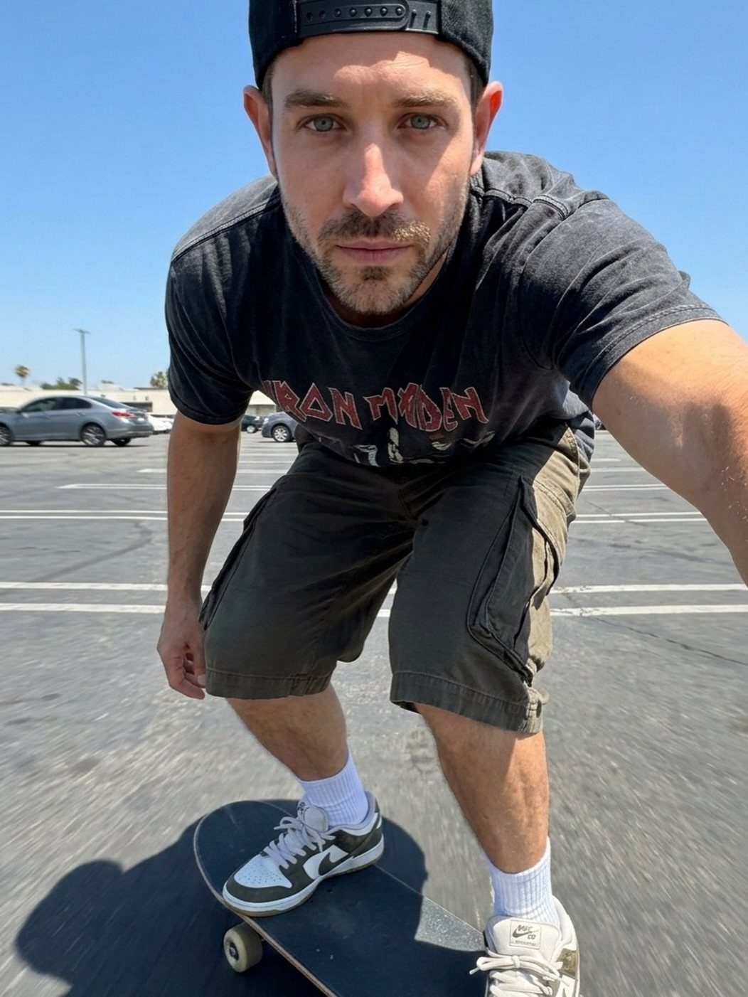 Man in graphic tee and cargo shorts standing on a skateboard in a parking lot.