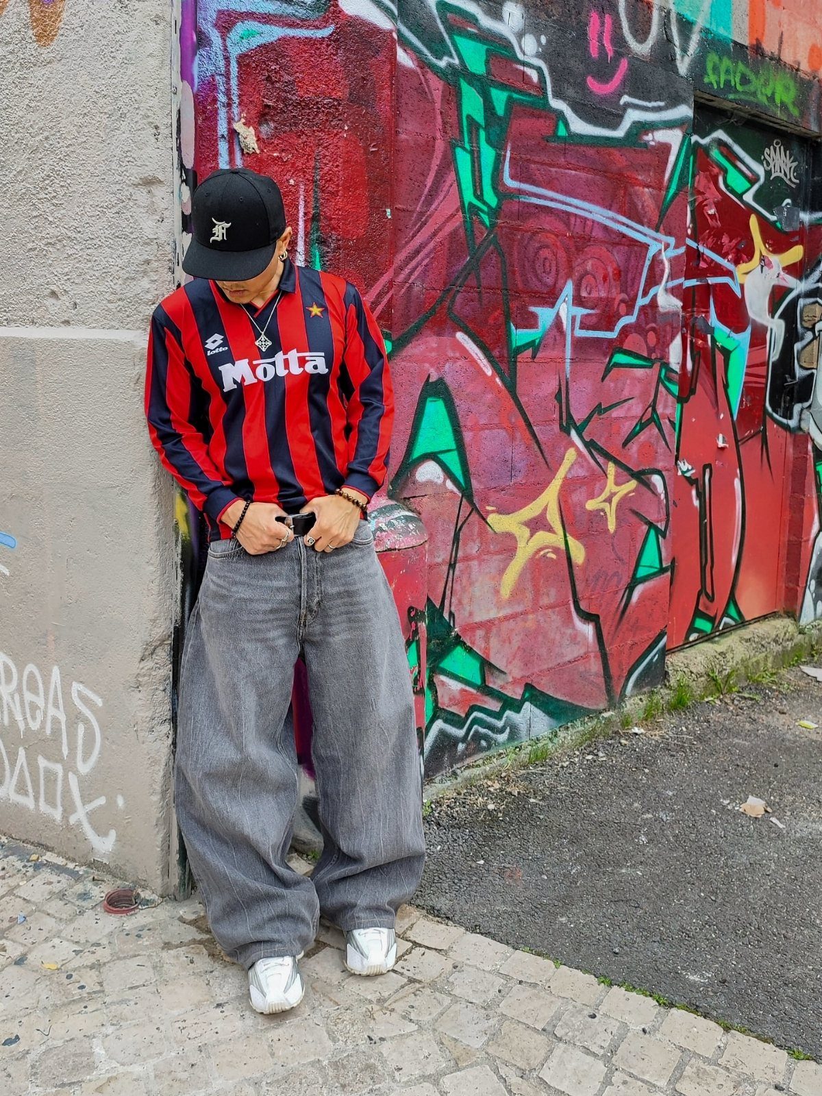 Man in baggy grey jeans and a striped jersey standing against a colorful graffiti wall.