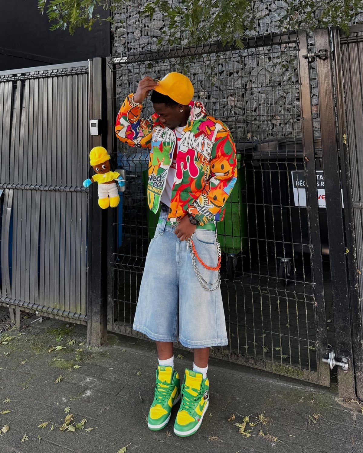 A person posing in a colorful patterned jacket, yellow beanie, denim shorts, and lime green sneakers.