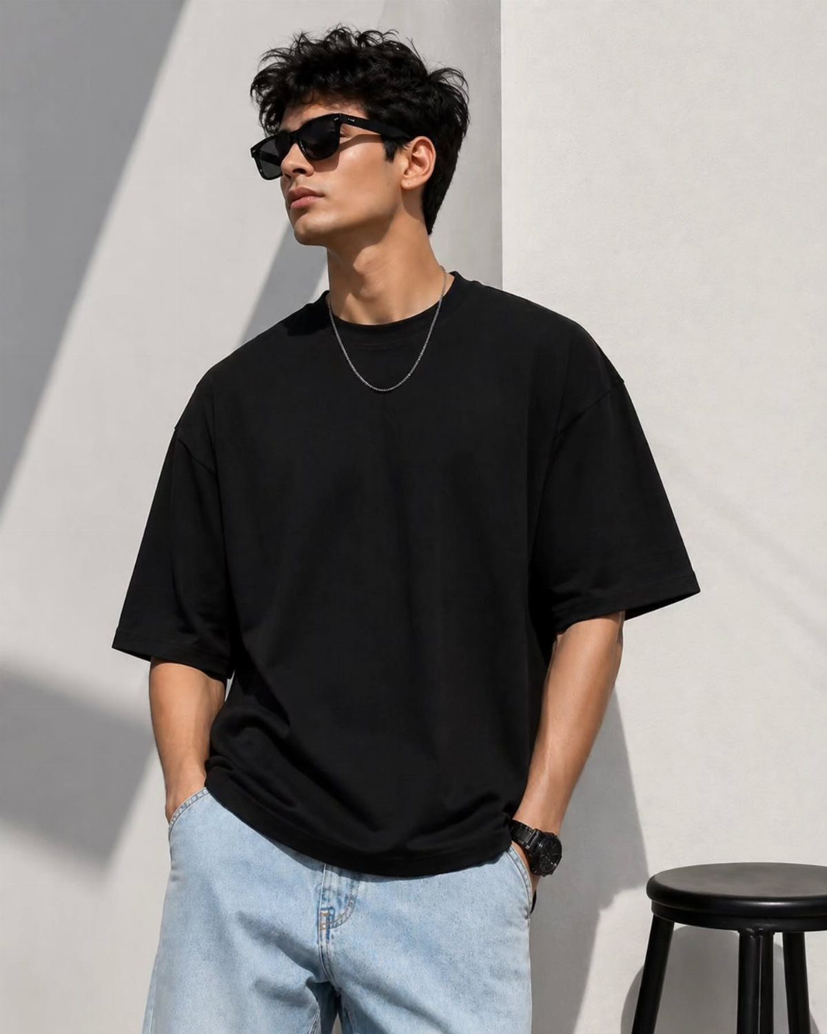 A man wearing a black oversized t-shirt, sunglasses, and light blue jeans standing against a white wall.