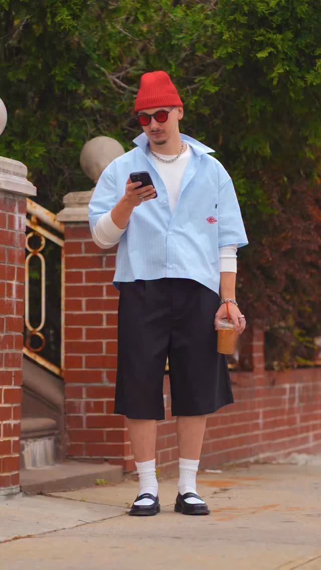 A man in a light blue striped shirt, red beanie, and black shorts stands on a sidewalk using his phone.