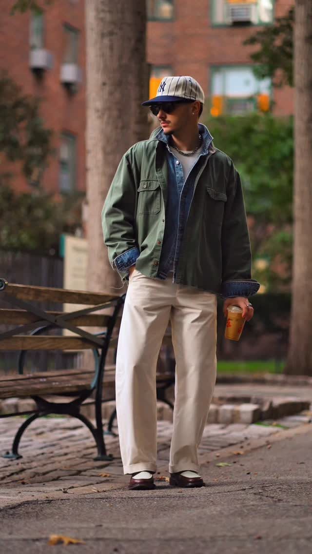 A man wearing a green jacket, denim shirt, and cream trousers standing in a park.