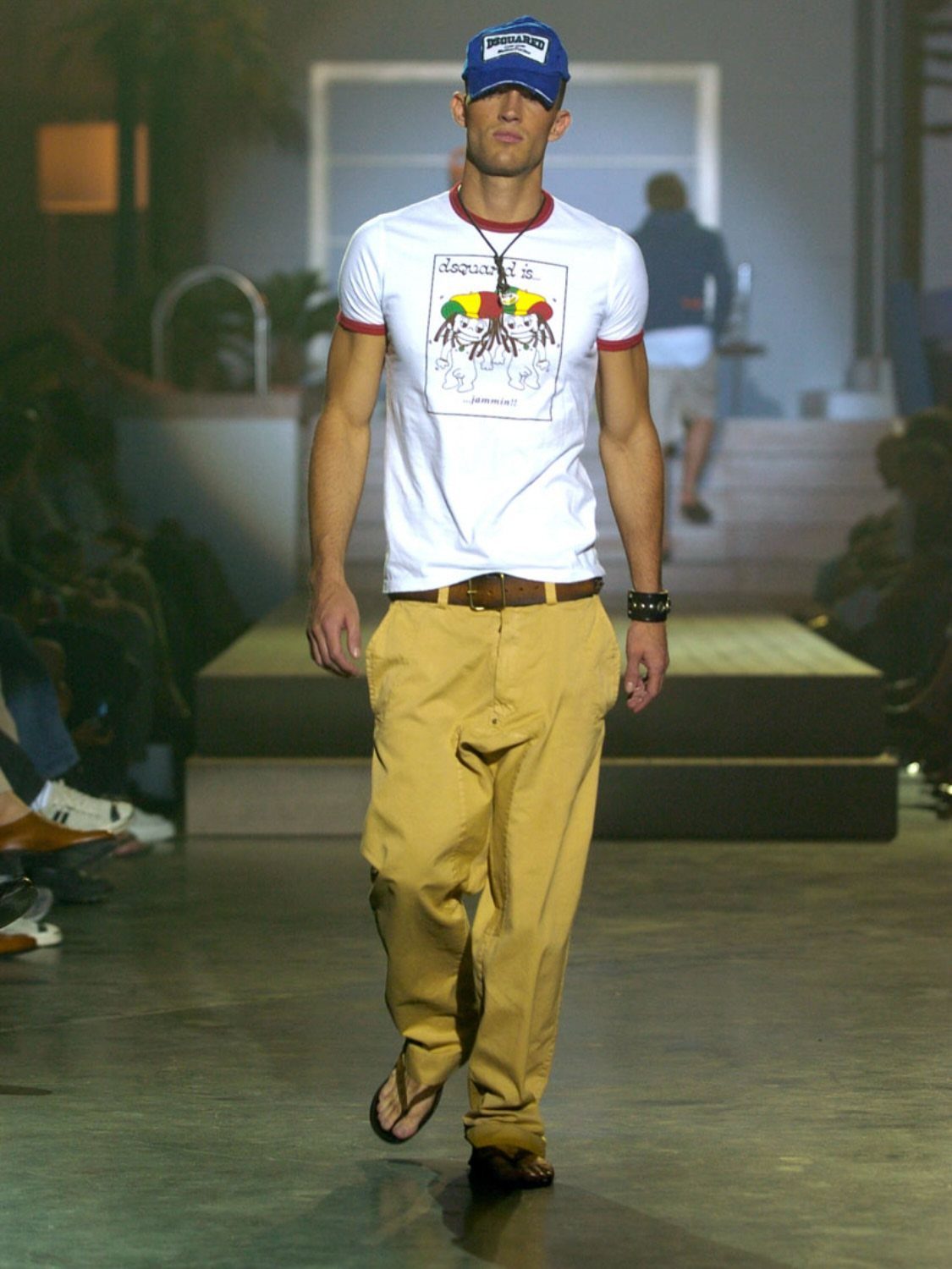 Male model walking a runway in a white graphic t-shirt, tan trousers, and a blue cap.