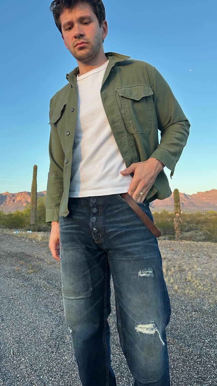 A man wearing an olive shirt, white t-shirt, and distressed blue jeans standing in a desert.