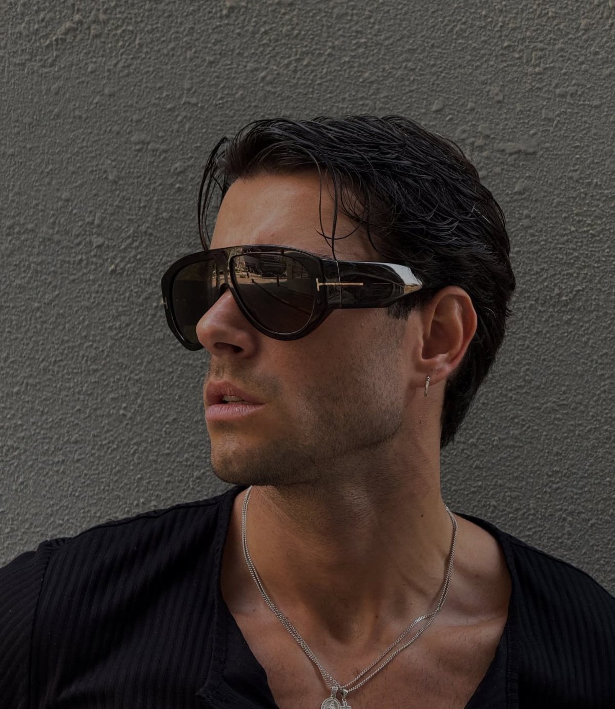 A man wearing large black oversized sunglasses and a silver necklace against a grey wall.