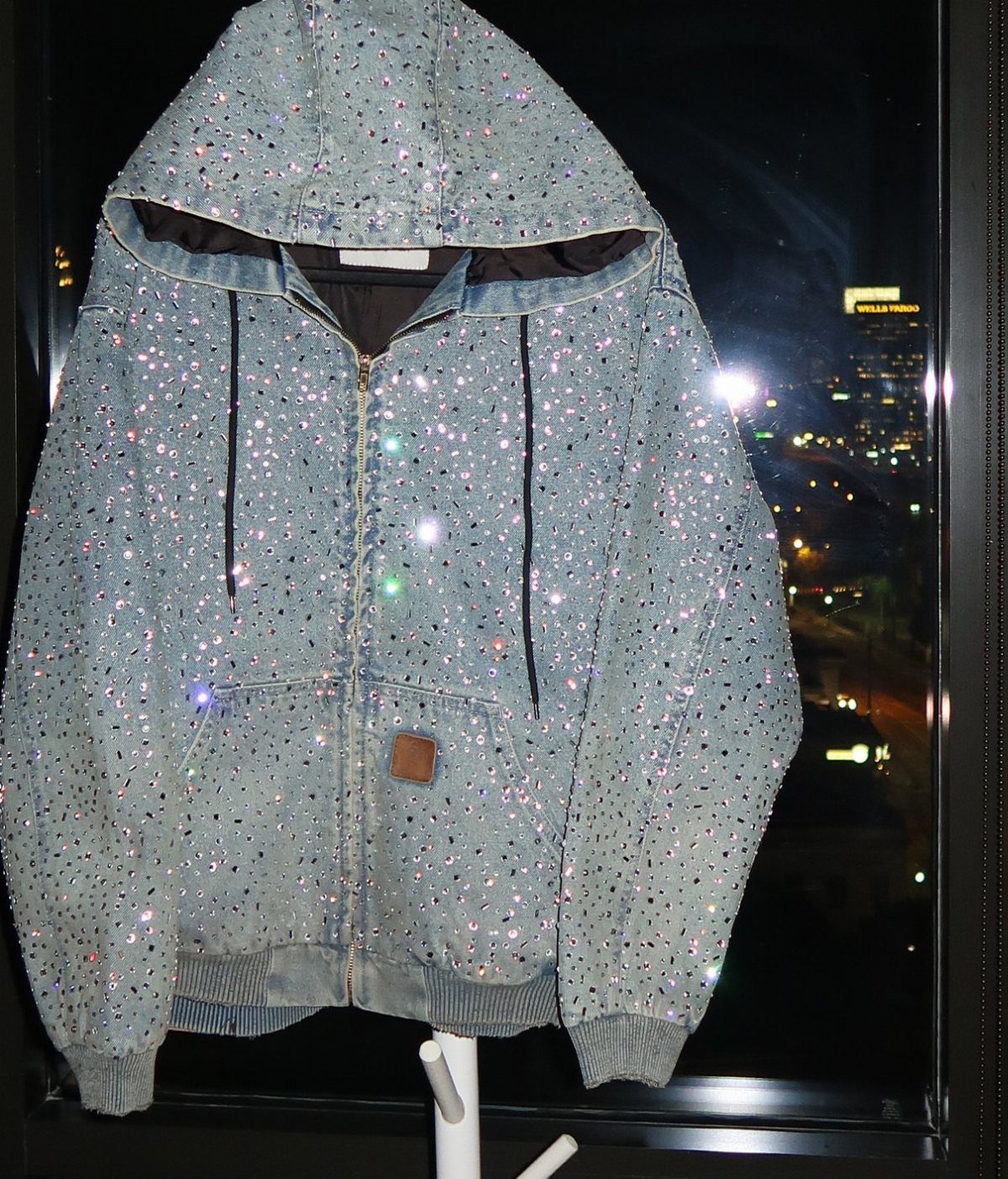 Light blue denim hooded jacket embellished with many small, sparkling rhinestones against a dark window.