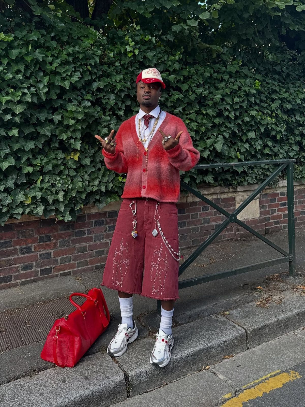 Man in vibrant red gradient cardigan and matching shorts posing against green ivy.