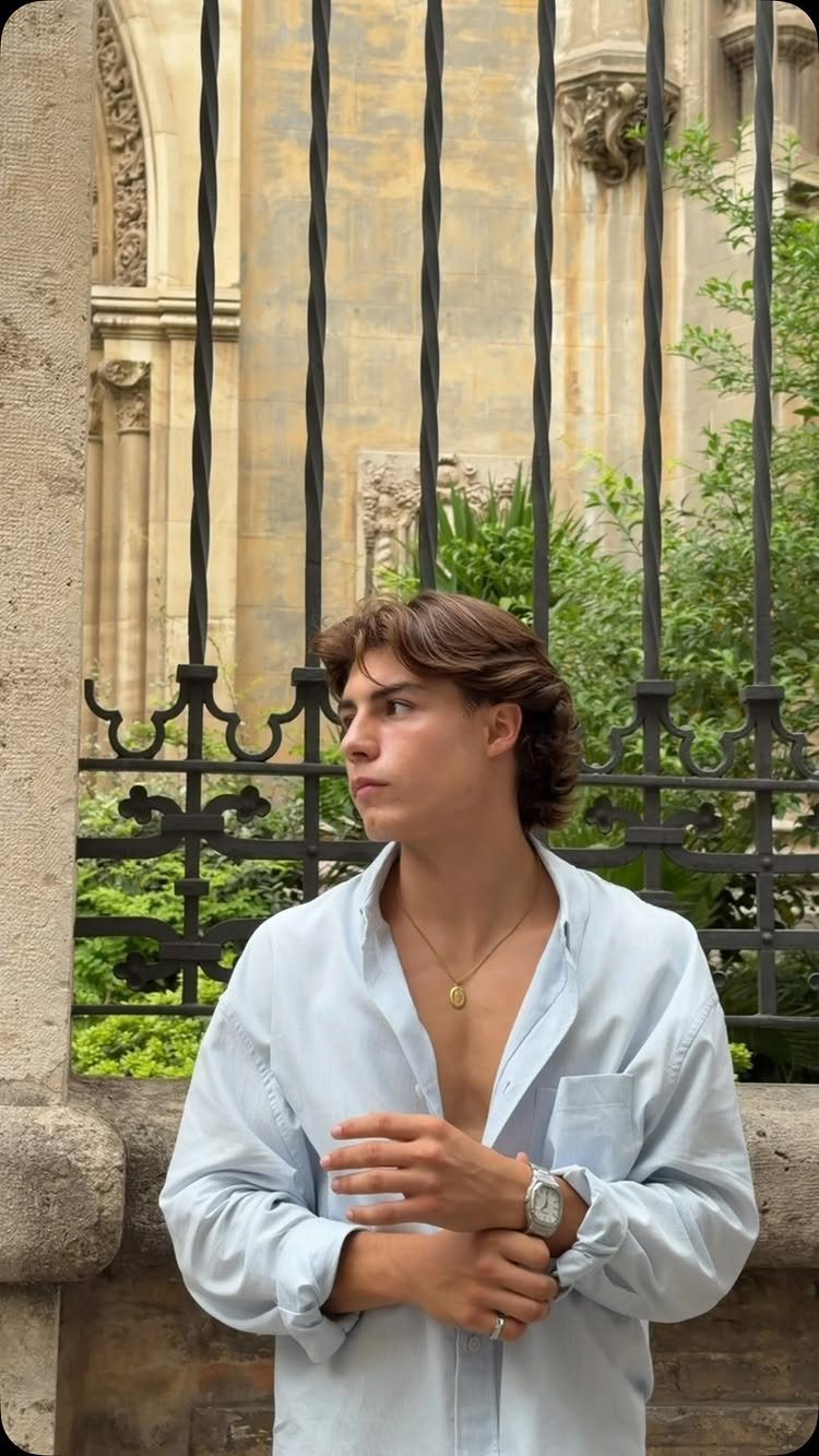 Young man wearing an open light blue shirt and gold necklace standing by a fence.